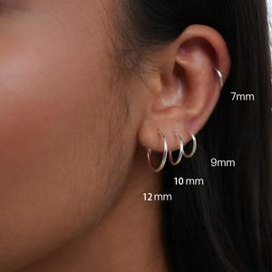 Puede incluir: Un conjunto de cuatro pendientes de aro plateados usados en una sola oreja, etiquetados con los tamaños 7mm, 9mm, 10mm y 12mm. Los aros son finos, circulares y metálicos, dispuestos en una progresión vertical desde el cartílago hasta el lóbulo.