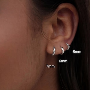 Könnte beinhalten: Nahaufnahme eines Ohrs mit drei silbernen Creolen. Die Ohrringe haben einen Durchmesser von 5 mm, 6 mm und 7 mm. Die Ohrringe glänzen und reflektieren das Licht. Das Ohr ist vor einem unscharfen Hintergrund.