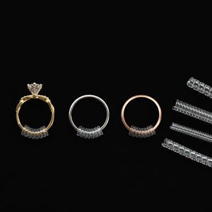 Puede incluir: Tres anillos en oro, plata y oro rosa, cada uno equipado con un ajustador de tamaño de anillo de plástico transparente. Se muestran varios ajustadores adicionales a la derecha. Fondo negro.