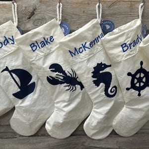 May include: A row of white Christmas stockings with navy blue accents, each personalized with a name and nautical-themed design. Designs include a paw print, sailboat, lobster, seahorse, ship's wheel, and another paw print.