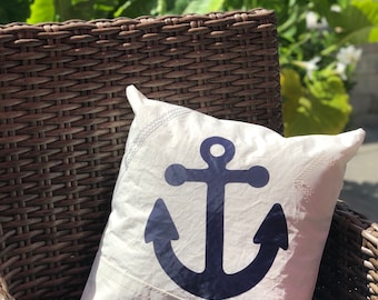 May include: A square white throw pillow featuring a large, dark navy blue anchor graphic centered on the front. The pillow fabric has a textured, sail-like appearance with visible zigzag stitching along the edges and corners.