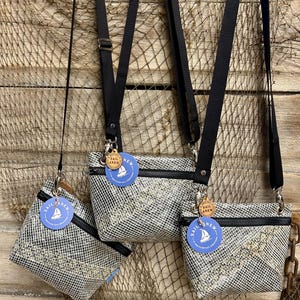 May include: Three crossbody bags made of white and black grid-patterned material with black zippers and adjustable black nylon straps. Each bag features a round blue tag with a white sailboat icon and the text SAIL ANEW, plus a small wooden tag.
