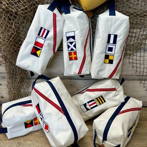 May include: White rectangular pouches with navy blue zippers and handles, featuring colorful nautical flag designs. The pouches have red zigzag stitching and are arranged on a wooden surface with fishing net and yellow buoys in the background.