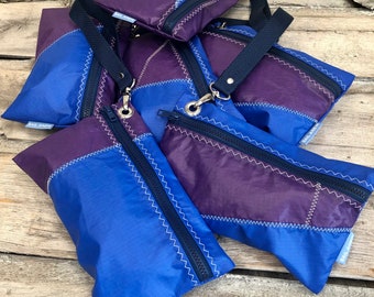 May include: Five blue and purple zippered pouches with a navy blue strap. The pouches are made from recycled sailcloth and have the text "Sail Anew" printed on them.