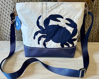 May include: White and navy blue tote bag with a crab design. The bag features a navy blue crab graphic on the front, a navy blue bottom, and a blue adjustable shoulder strap. The bag is made of a sailcloth material.