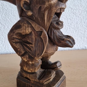 Könnte beinhalten: Handgeschnitzte Holzfigur eines Zwergs mit übertriebenem Gesichtsausdruck. Der Zwerg hat eine große Nase, einen offenen Mund und einen langen Bart. Die Figur ist braun und steht auf einem rechteckigen Sockel.