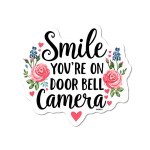 Op de afbeelding: Een witte sticker met de tekst "Smile, You're on Door Bell Camera" in zwart schrift. Het ontwerp bevat roze rozen, blauwe bloemen en kleine rode hartjes.