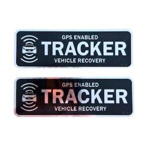 Könnte beinhalten: Zwei schwarze und silberne rechteckige Aufkleber. Jeder Aufkleber trägt die weißen Worte "GPS ENABLED TRACKER VEHICLE RECOVERY". Ein Autosymbol und Signalwellen sind ebenfalls vorhanden.