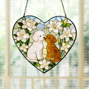 Può includere: Un acchiappasole in vetro colorato a forma di cuore con un cane bianco e un cane marrone seduti insieme, circondati da fiori di giglio bianchi e foglie verdi. Il pezzo è incorniciato in metallo nero e appeso a una catena nera.