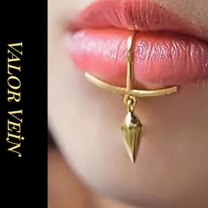 Könnte beinhalten: Nahaufnahme eines goldenen Lippenrings mit einem geometrischen Anhänger. Der Schmuck ist an der Unterlippe befestigt. Der Text "VALOR VEIN JEWELRY" befindet sich auf schwarzem Hintergrund.