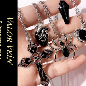 Puede incluir: Una colección de collares plateados con colgantes góticos, incluyendo una cruz, un corazón, una araña, un ataúd y un corazón anatómico. Los colgantes son negros y plateados. El texto "VALOR VEIN JEWELRY" está a la izquierda.