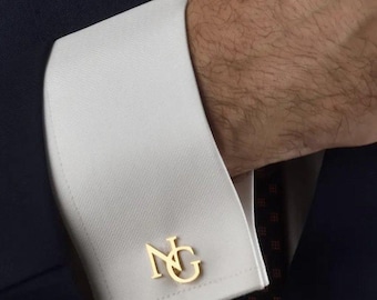 Può includere: Un gemello in metallo dorato con le lettere intrecciate NG, fissato attraverso le asole di una manica di camicia bianca con polsino alla francese. La camicia è indossata sotto un blazer blu scuro con un sottile motivo testurizzato.