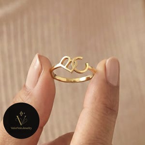 May include: A gold-toned ring featuring two interconnected letters, B and C, forming a stylized design. The band is thin and polished, showcasing a metallic finish. A circular black logo with the text ValorVeinJewelry is visible in the corner.