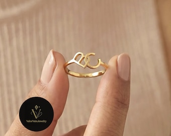 May include: A gold-toned ring featuring two interconnected letters, B and C, forming a stylized design. The band is thin and polished, showcasing a metallic finish. A circular black logo with the text ValorVeinJewelry is visible in the corner.