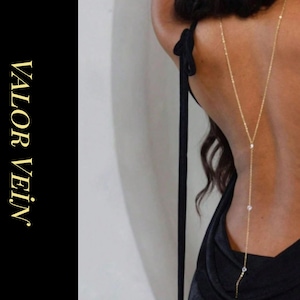 Puede incluir: Un collar dorado para la espalda con tres pequeñas piedras preciosas transparentes. El collar cuelga por la espalda de una persona que lleva una parte superior negra. Las palabras "VALOR VEIN JEWELRY" están impresas en oro sobre un fondo negro.