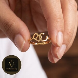 May include: A gold-toned ring featuring a polished metal band with a decorative centerpiece shaped like an interlocking heart and the letter C. The design is crafted from a smooth, reflective material with a consistent metallic finish throughout.