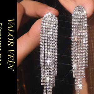 Puede incluir: Pendientes colgantes plateados con múltiples hilos de brillantes pedrería. Los pendientes se sostienen cerca de la oreja, mostrando su diseño. El texto "VALOR VEIN JEWELRY" está en el lado izquierdo de la imagen.