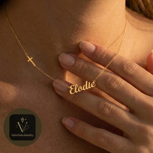 May include: A gold-toned necklace featuring a delicate chain with a small cross charm positioned to the left and a cursive nameplate pendant that reads Elodie in the center. The metal has a polished finish and rests against the skin.