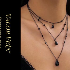 Puede incluir: Un collar negro de tres capas con colgantes en forma de lágrima y pequeñas cuentas. El collar está hecho de cadenas delicadas y se muestra en el cuello de una persona. La imagen también incluye el texto "VALOR VEIN JEWELRY" en letras doradas.