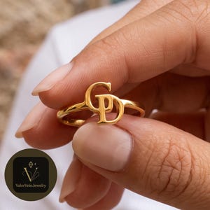 May include: A gold-toned ring featuring a polished finish with two interlocking capital letters, G and D, centered on the band. The letters are connected in a stylized design, creating a singular focal point on the thin, smooth metal band.