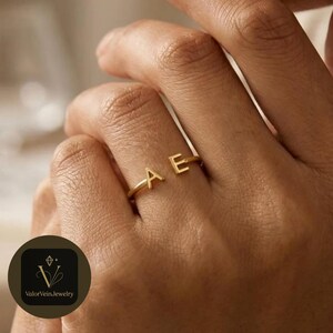 May include: A gold-toned open ring featuring two capital letters, A and E, positioned at the ends of the band. The ring has a polished finish and a simple, thin metal construction that wraps partially around the finger.