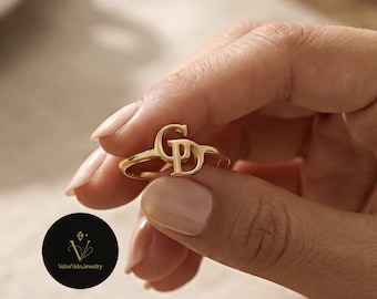 May include: A gold-toned ring featuring two interlocking capital letters, G and D, as the central focal point. The letters are connected in a continuous design, forming the main decorative element of the band. The ring has a polished finish.