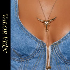 Könnte beinhalten: Eine goldfarbene Halskette mit einem Bullenschädel-Anhänger. Der Anhänger ist mit Hörnern und Gesichtszügen versehen. Die Halskette wird über einem Jeans-Top getragen. Die Worte "VALOR VEIN JEWELRY" sind vertikal gedruckt.