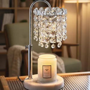 May include: A decorative table lamp with a chrome finish and a crystal shade. The lamp features a round marble base and a jar candle with a label that reads "Fleurgas". The candle is lit, casting a warm glow.