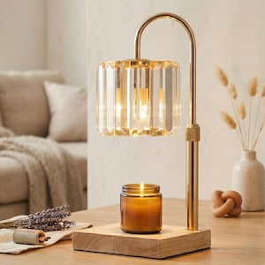 May include: A gold-toned lamp with a clear, faceted glass shade illuminates a wooden base. A lit candle in a glass jar sits on the base. The lamp's curved arm and base are made of gold-colored metal. A vase with dried plants and a decorative knot are in the background.