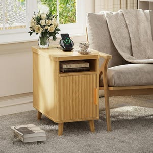May include: A light wood-tone bedside table with a single door and open shelf. A wireless charger with a phone sits on top, next to a teacup and a vase of flowers. The table is on a gray carpet, near a chair.