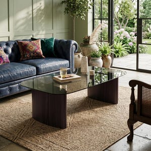 May include: A coffee table with a dark, smoked glass top and two curved, dark wood bases with vertical fluting. The table is rectangular with rounded corners and sits on a textured, woven rug. A small book and a glass sit on the table surface.