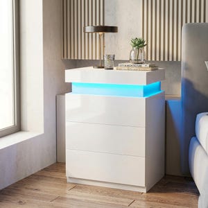 May include: A modern white nightstand with three drawers and a top shelf. The top shelf has a built-in blue LED light. A small lamp, a glass, a succulent, and books are on the top. The nightstand is next to a bed.