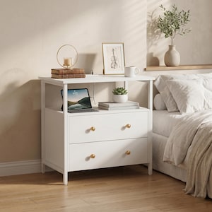 May include: A white nightstand with two drawers and gold-colored knobs. The top shelf holds a framed picture, a mug, a small succulent, and a tablet. A stack of books and a candle holder are also on the top shelf. The nightstand is next to a bed.