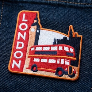 May include: Embroidered patch featuring a red double-decker bus, Big Ben, and London skyline. The patch has an orange border and the word "LONDON" in white vertical letters on a red background. The patch is set against a dark blue denim background.