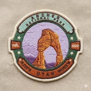 May include: Embroidered patch featuring the Arches National Park in Utah. The patch has a circular design with the park name and "est. 1929." The image depicts a sandstone arch against a purple and blue sky.