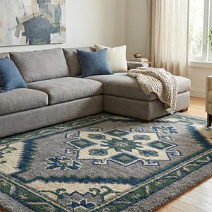 May include: A grey corner sofa with blue and beige cushions is placed on a patterned rug. The rug has a central geometric design in blue, green, and cream. A beige armchair and a window with plants are also visible.