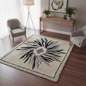 May include: A rectangular area rug with a floral design in shades of gray and black on a cream background. The rug is bordered with a black trim and is placed on a dark wood floor. Two gray armchairs are on either side of the rug.