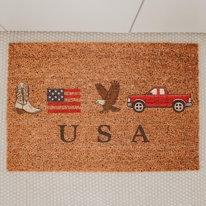 May include: A rectangular doormat with a brown coir surface. The mat features a cowboy boot, American flag, eagle, and red pickup truck graphic. The word "USA" is printed in brown block letters below the graphics.