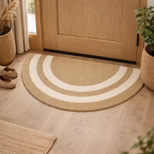 May include: A half-circle beige and white striped doormat sits on a light wood floor in front of a wooden door. The mat has two white stripes. A small rectangular rug is in the lower left corner. Wicker baskets with plants are on either side.