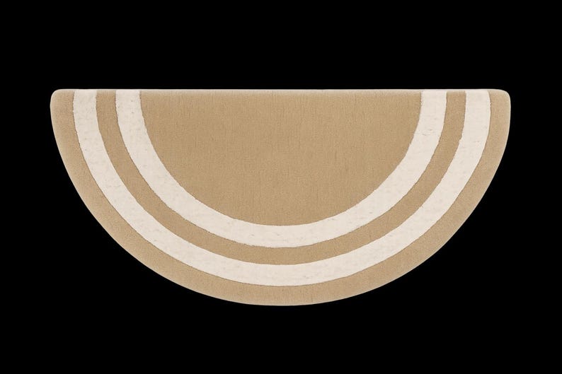 May include: A half-circle rug in a neutral tan color with three off-white curved stripes. The rug has a soft, textured appearance and is set against a black background. The design is simple and modern.
