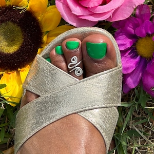 May include: A foot wearing a gold metallic sandal with a silver toe ring. The toenails are painted bright green. The background includes a yellow sunflower, a pink rose, and purple flowers.
