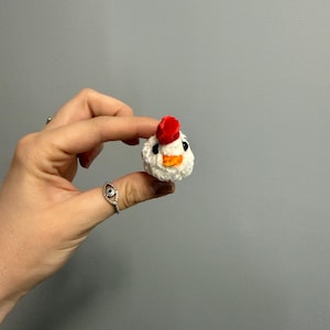 May include: A small, handmade, white crochet chicken head with a red comb, orange beak, and black eyes. The chicken is held up against a gray background.