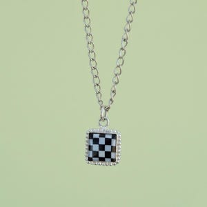 May include: Silver-toned necklace with a square pendant featuring a black and white checkered pattern. The pendant is framed with a beaded border. The chain is a delicate link style, and the background is a soft green.