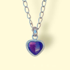 Puede incluir: Collar plateado con un colgante en forma de corazón. El colgante tiene un centro morado y azul, enmarcado por un borde plateado en forma de cuerda. La cadena está hecha de eslabones grandes y ovalados.