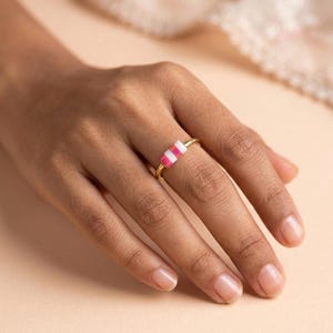 May include: A gold ring featuring a rectangular design with alternating pink and white stripes. The ring is on a finger, with a soft, neutral background.