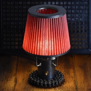Peut inclure: Une lampe de table dotée d'un abat-jour conique plissé rouge avec un bord à carreaux noir et gris. La base est constituée d'un élément de tuyau cylindrique noir fixé à un assemblage circulaire de maillons de chaîne, avec un petit interrupteur doré.