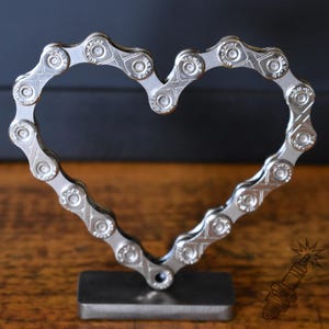 Könnte beinhalten: Eine herzförmige Skulptur aus einer silberfarbenen Fahrradkette aus Metall, bei der auf jedem Glied der Schriftzug MSN-SPORT eingeprägt ist. Die Kette ist auf einem rechteckigen, dunkelgrauen Metallsockel für einen stabilen Stand montiert.