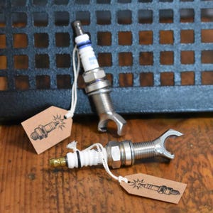 Könnte beinhalten: Zwei Zündkerzen, die an der Basis mit einem U-förmigen Metallaufsatz modifiziert wurden. Jede Zündkerze hat einen weißen Keramikisolator, einen silbernen Gewindekörper und ein braunes Papieretikett, das mit einer weißen Schnur befestigt ist.