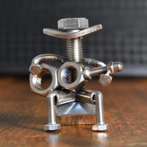 Könnte beinhalten: Handgefertigte Metallskulptur einer Person, die Gitarre spielt, aus Muttern, Schrauben und Unterlegscheiben gefertigt. Die Figur sitzt, mit einer Schraube als Hut und einer Gitarre aus Metallkomponenten. Die Skulptur steht auf einer Holzoberfläche.