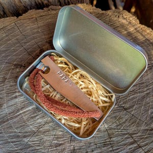 May include: A rectangular metal tin containing a copper-colored metal bottle opener with the word PAPA and a small heart symbol stamped on it. A textured, reddish-brown leather cord is attached to the opener, which rests on wood shavings.
